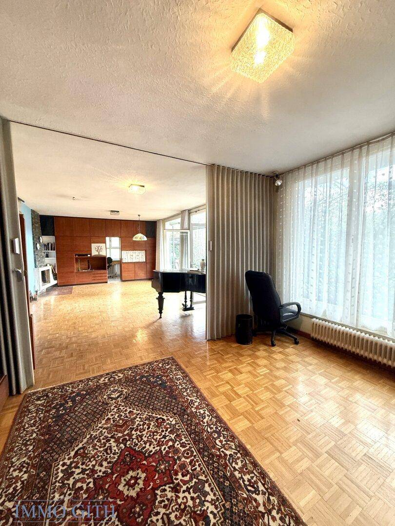 Weitläufiges Wohnzimmer mit Parkettboden, großen Fenstern und Blick in den angrenzenden Bereich.