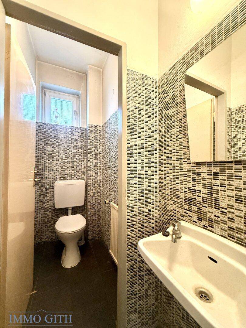 Separate Toilette mit Mosaikfliesen, Waschbecken und Fenster für Belüftung.