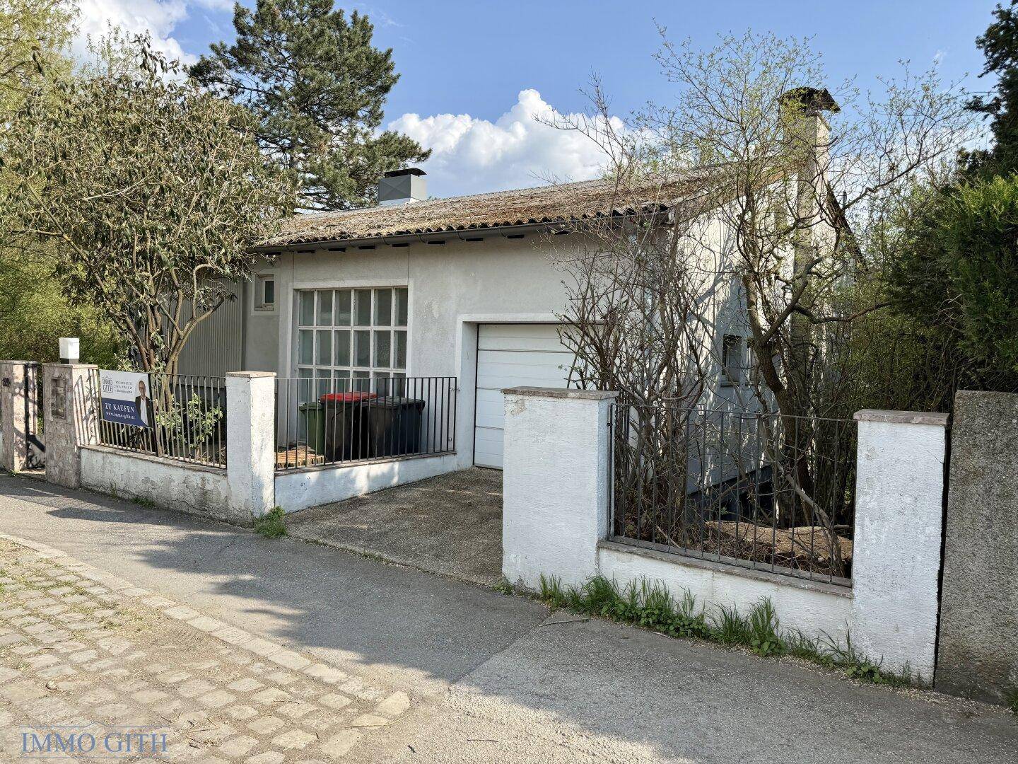 Hausfassade mit Garage, Einfahrt und Mülltonnenbereich, umgeben von Vegetation.