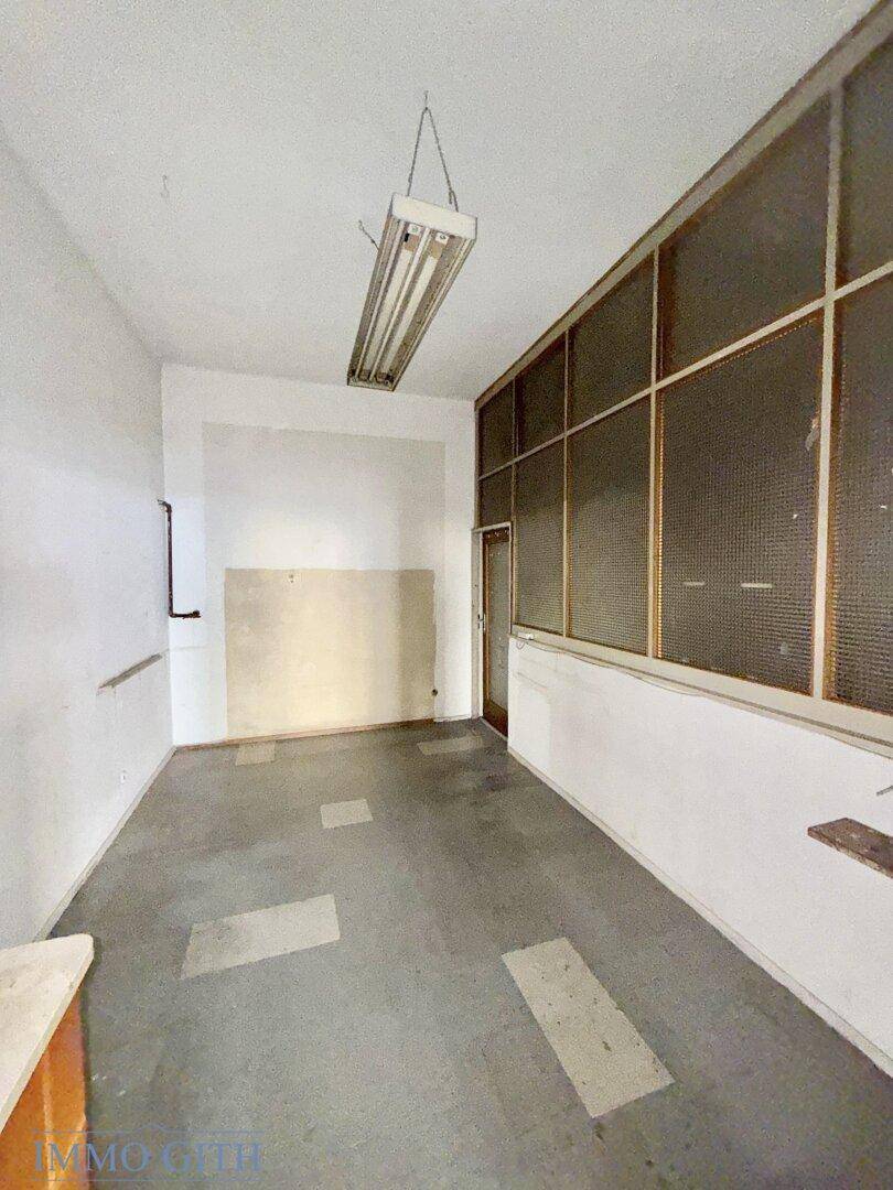 Leerer Raum mit weißen Wänden, grauem Boden und einem Fenster mit Gitter.