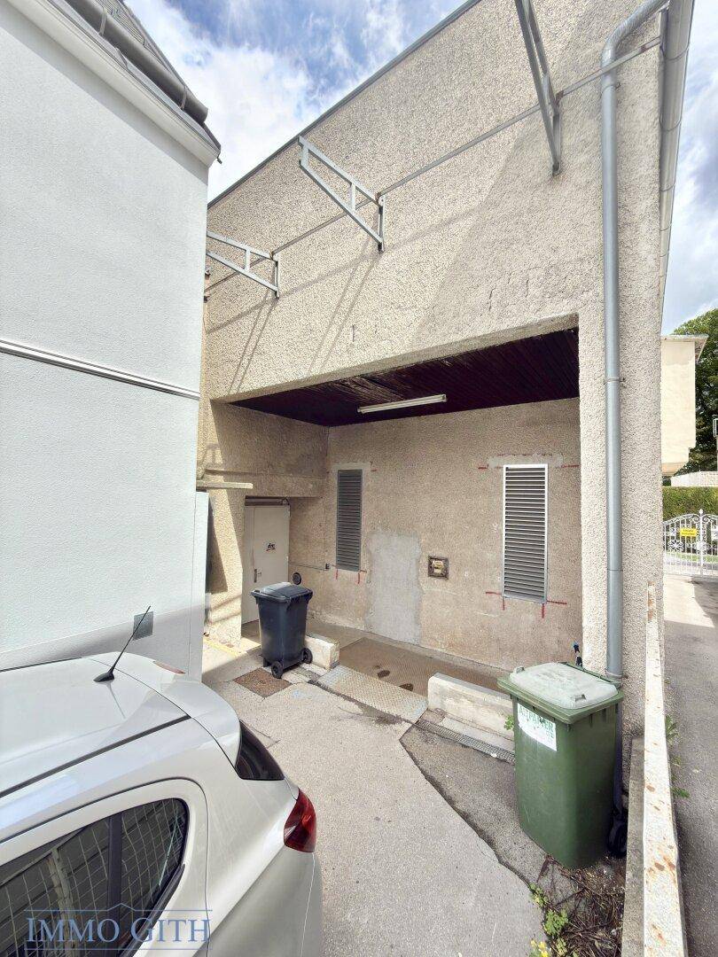 Rückansicht eines Gebäudes mit Zufahrt, Mülltonnen und Lüftungsschlitzen an der Fassade.