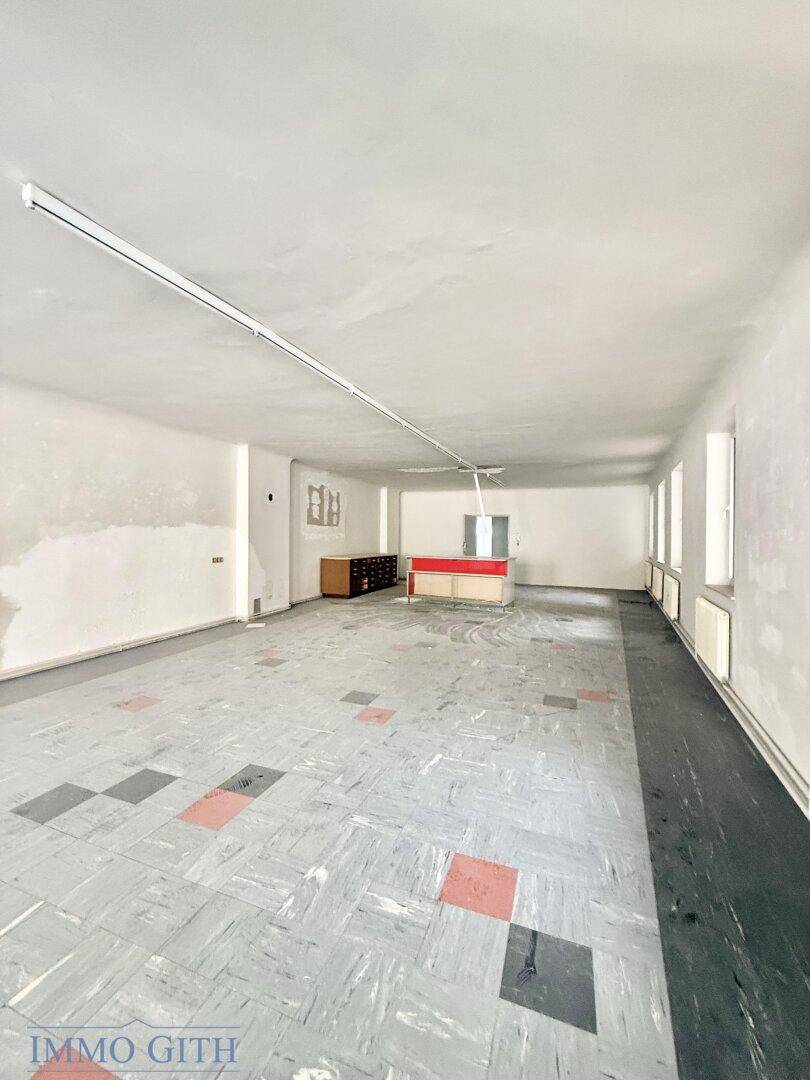 Weitläufiger Innenraum mit altem Bodenbelag und einer Theke im Hintergrund, unmöbliert.