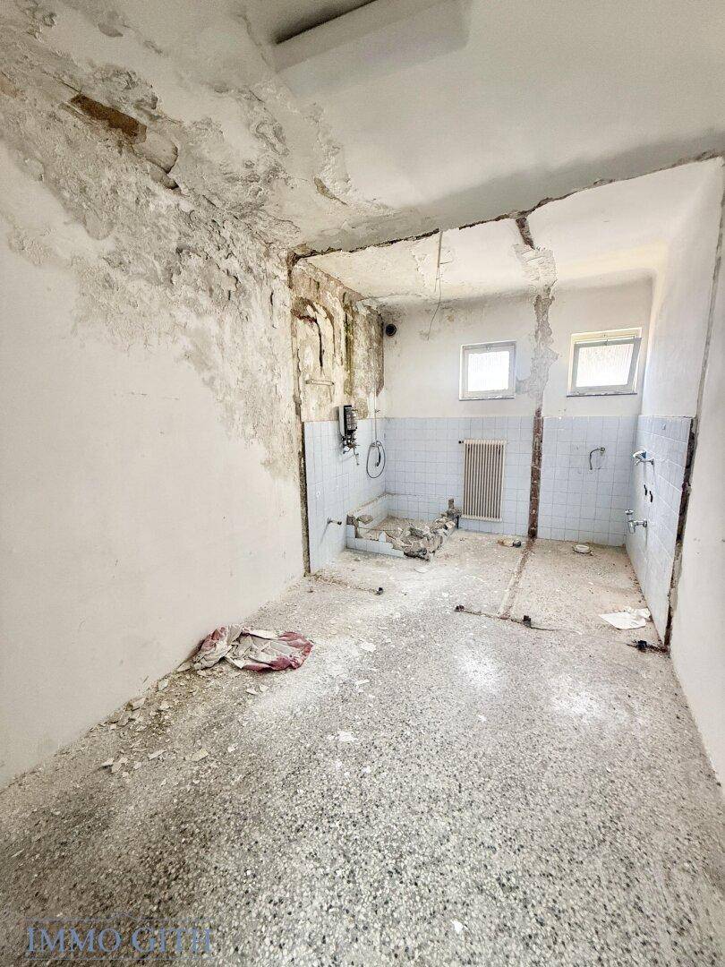 Renovierungsbedürftiges Badezimmer mit alten Fliesen und sichtbaren Wasseranschlüssen, zwei Fenstern.