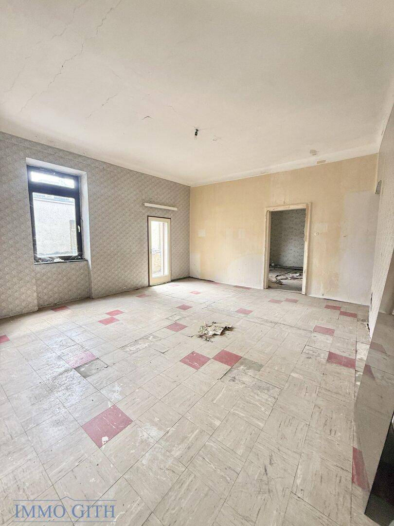 Geräumiger Raum mit altem Tapetenmuster, abgenutztem Boden und einem Fenster, renovierungsbedürftig.