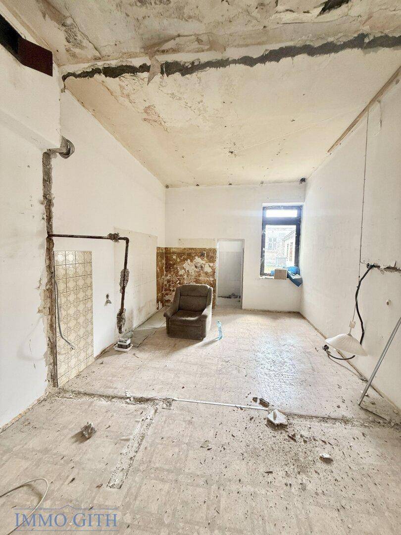 Renovierungsbedürftiger Wohnraum mit freiliegenden Rohren und einem alten Sessel, viel Potenzial.