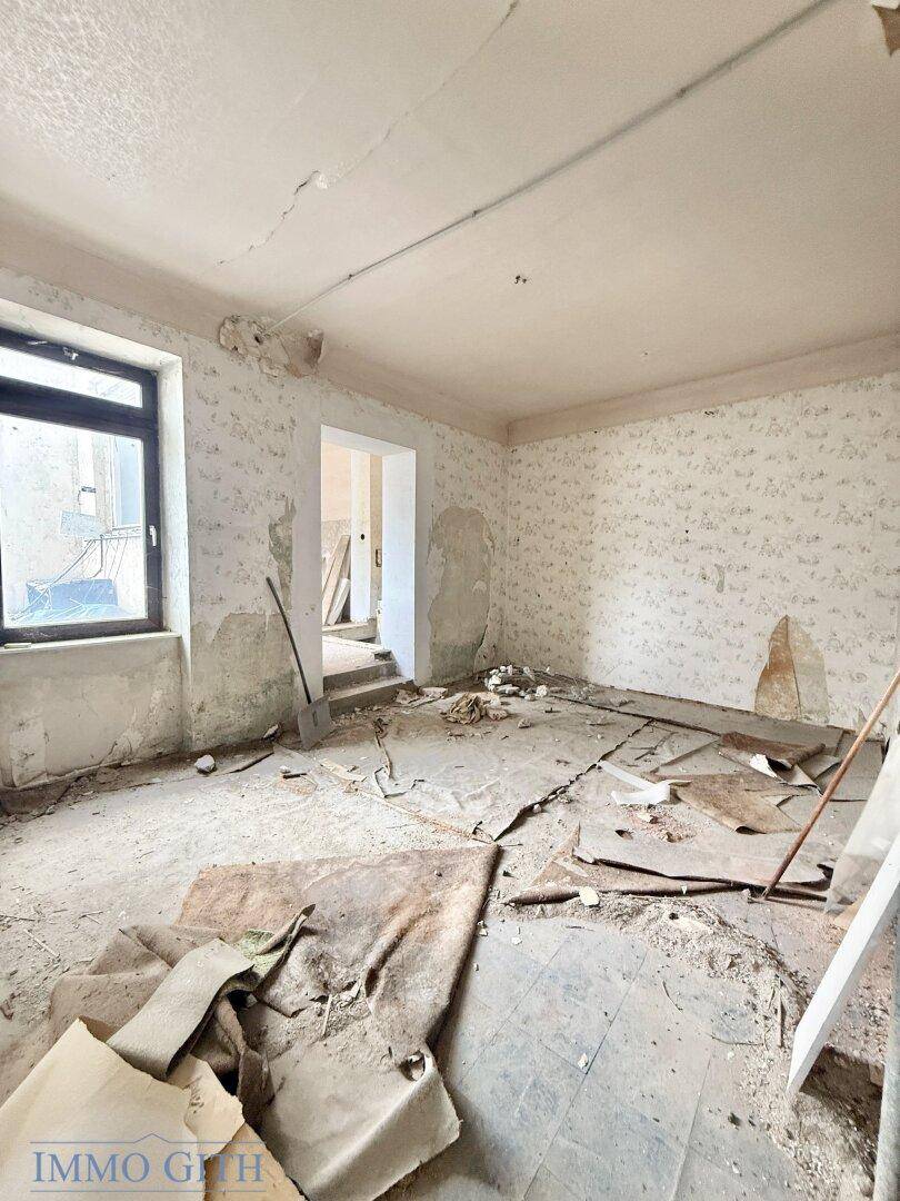 Großer Raum mit alter Tapete, abgenutztem Boden und Blick auf einen weiteren Raum, unrenoviert.