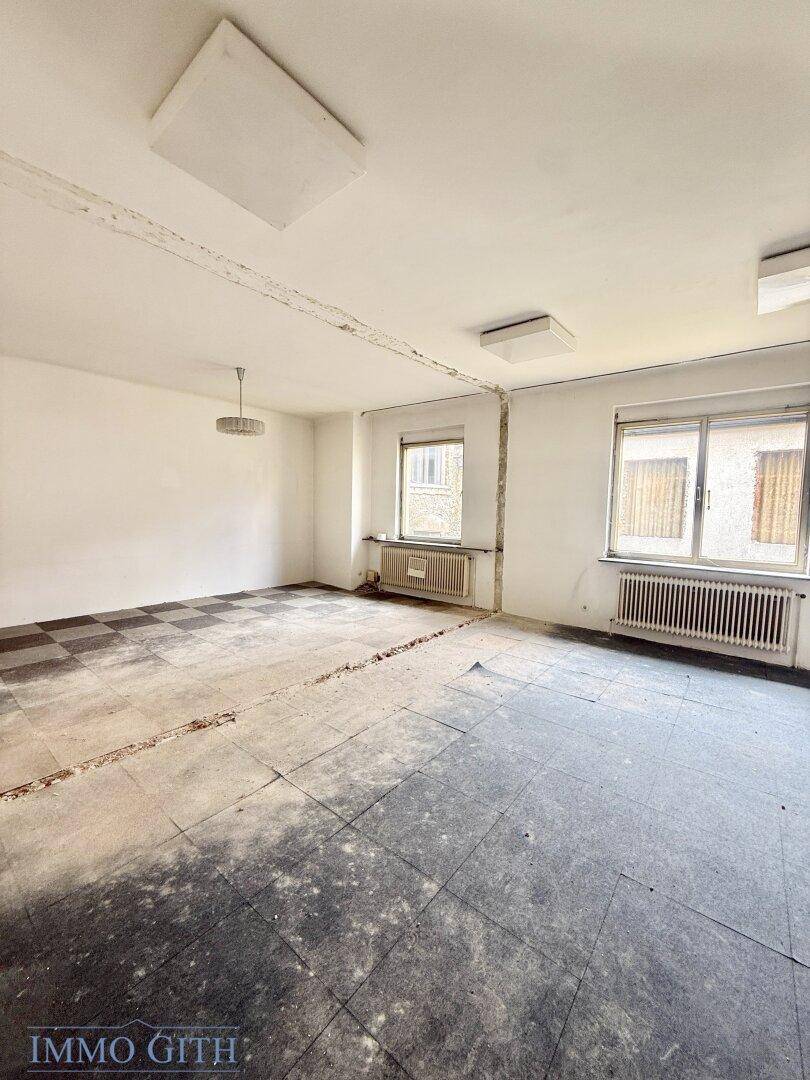 Geräumiger, unmöblierter Raum mit Fenstern und einem alten Bodenbelag, bereit zur Renovierung.