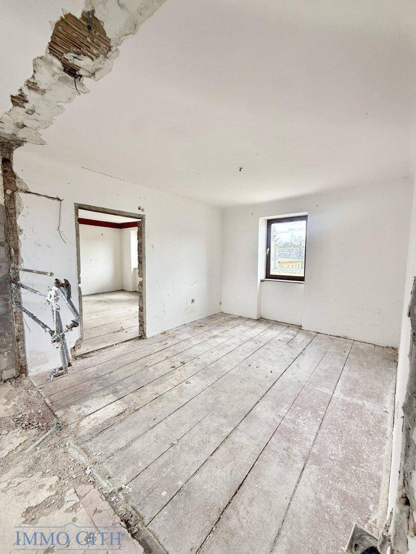 Renovierungsbedürftiger Raum mit freigelegten Holzdielen, weiß gestrichenen Wänden und einem Fenster.