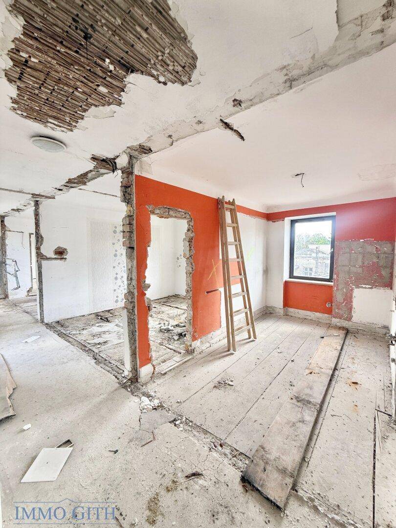 Innenraum im Rohbau mit sichtbaren Ziegelwänden, orangefarbenen Akzenten und einer Leiter.