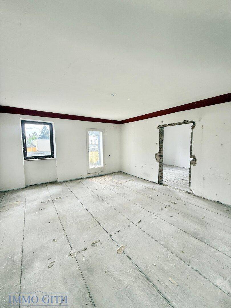 Renovierungsbedürftiger Innenraum mit freigelegten Holzdielen, weißen Wänden und einem Fenster.