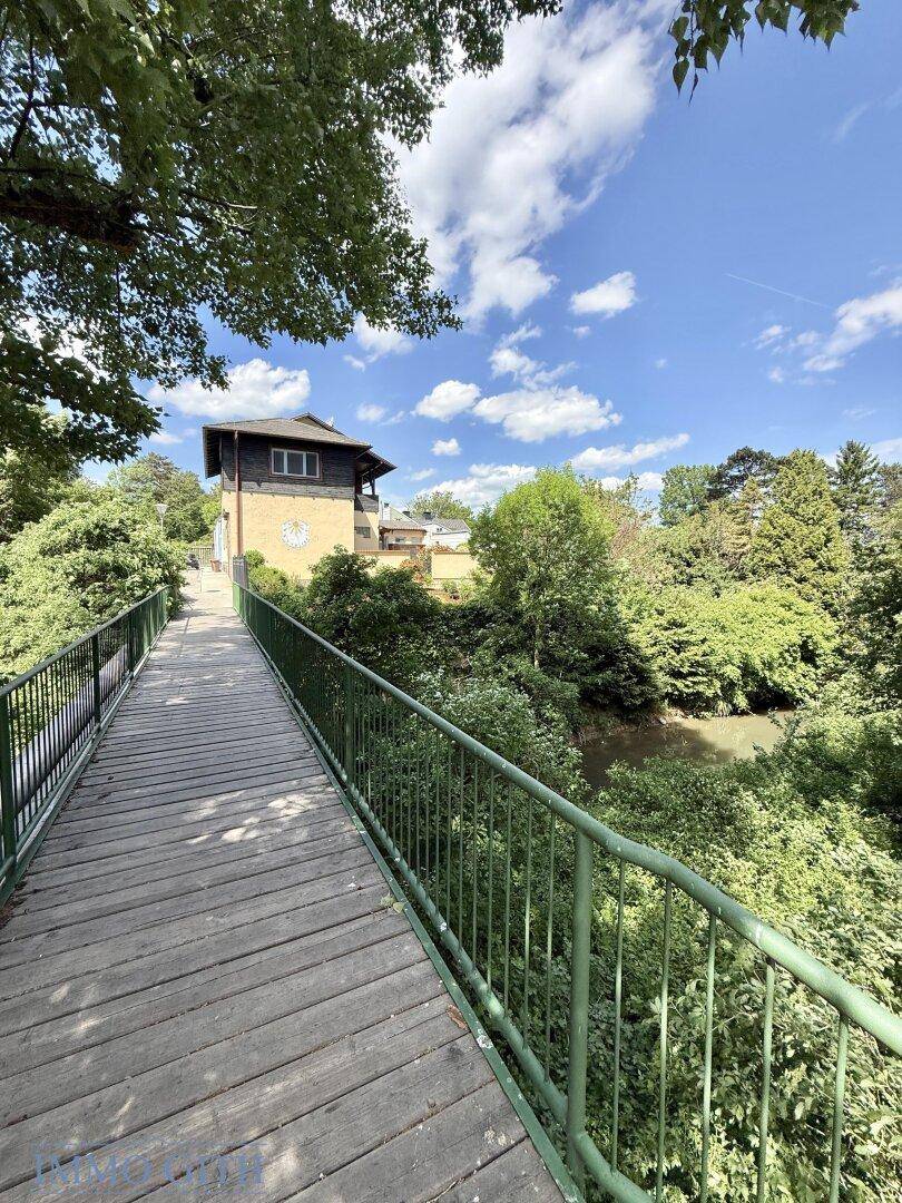 Holzbrücke über einen Fluss mit üppiger Vegetation und einem Gebäude im Hintergrund.