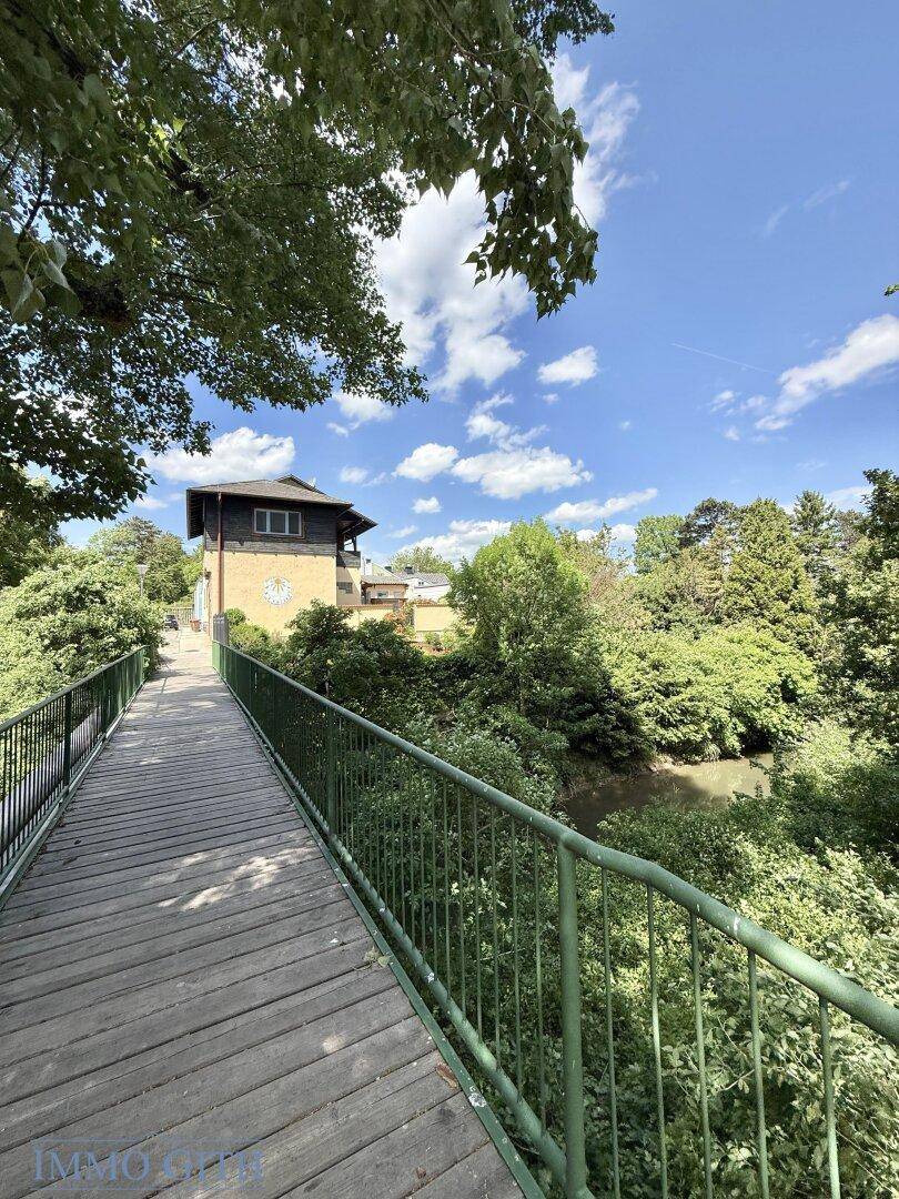 Holzbrücke über einen Fluss mit üppiger Vegetation und einem Gebäude im Hintergrund.