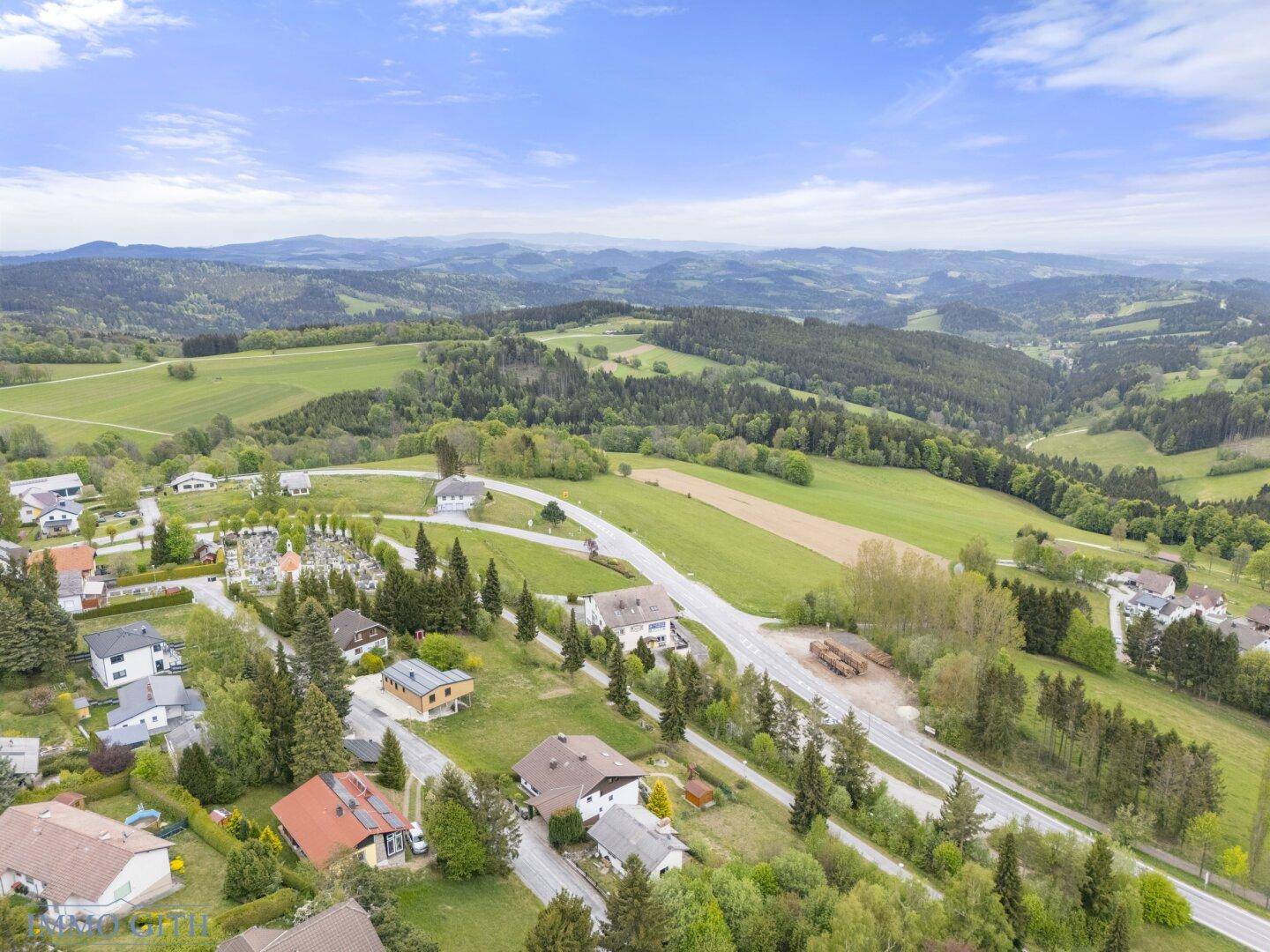 Weitläufige Landschaft mit grünen Hügeln, Wäldern und verstreuten Häusern unter blauem Himmel.