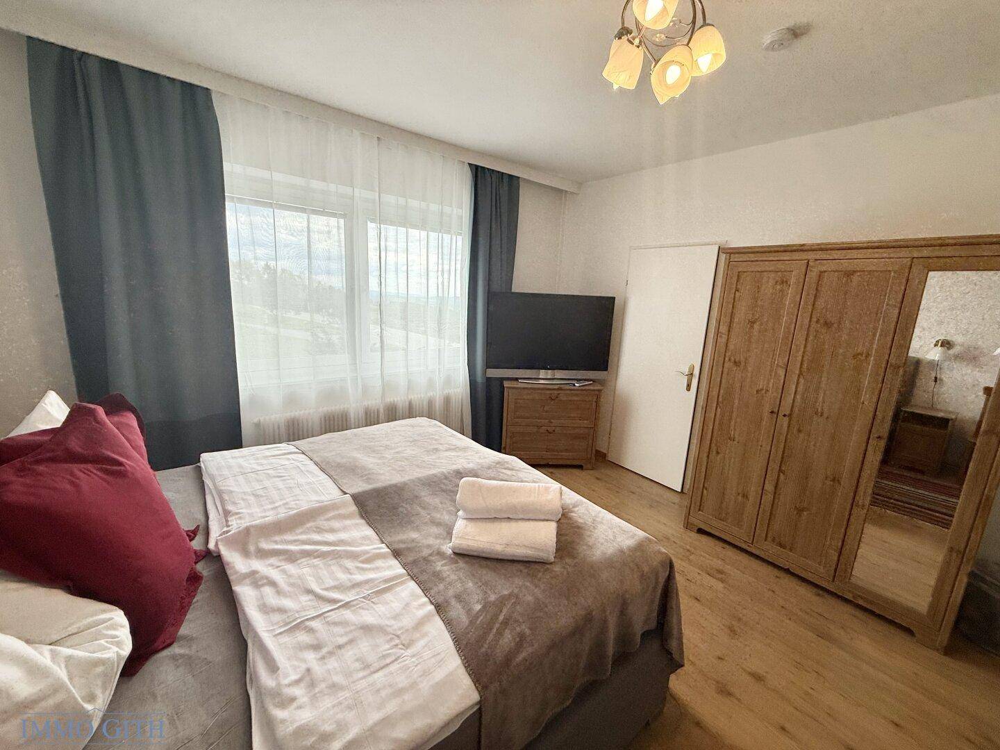 Schlafzimmer mit Doppelbett, Holzschrank und Fernseher, mit Blick nach draußen.