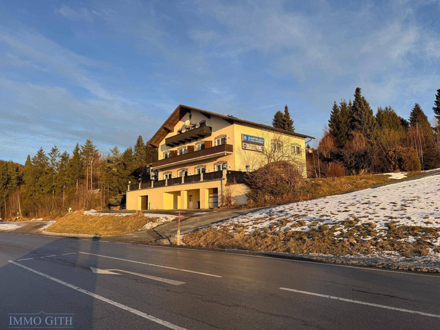Gebäude in ländlicher Umgebung mit verschneiten Hängen und Bäumen im Sonnenlicht.