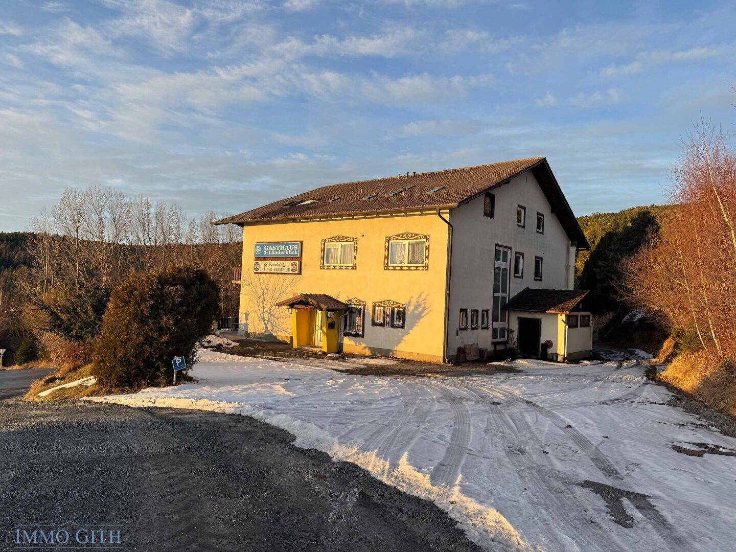 Gasthaus in ländlicher Umgebung mit verschneiter Zufahrt und Bäumen im Hintergrund.