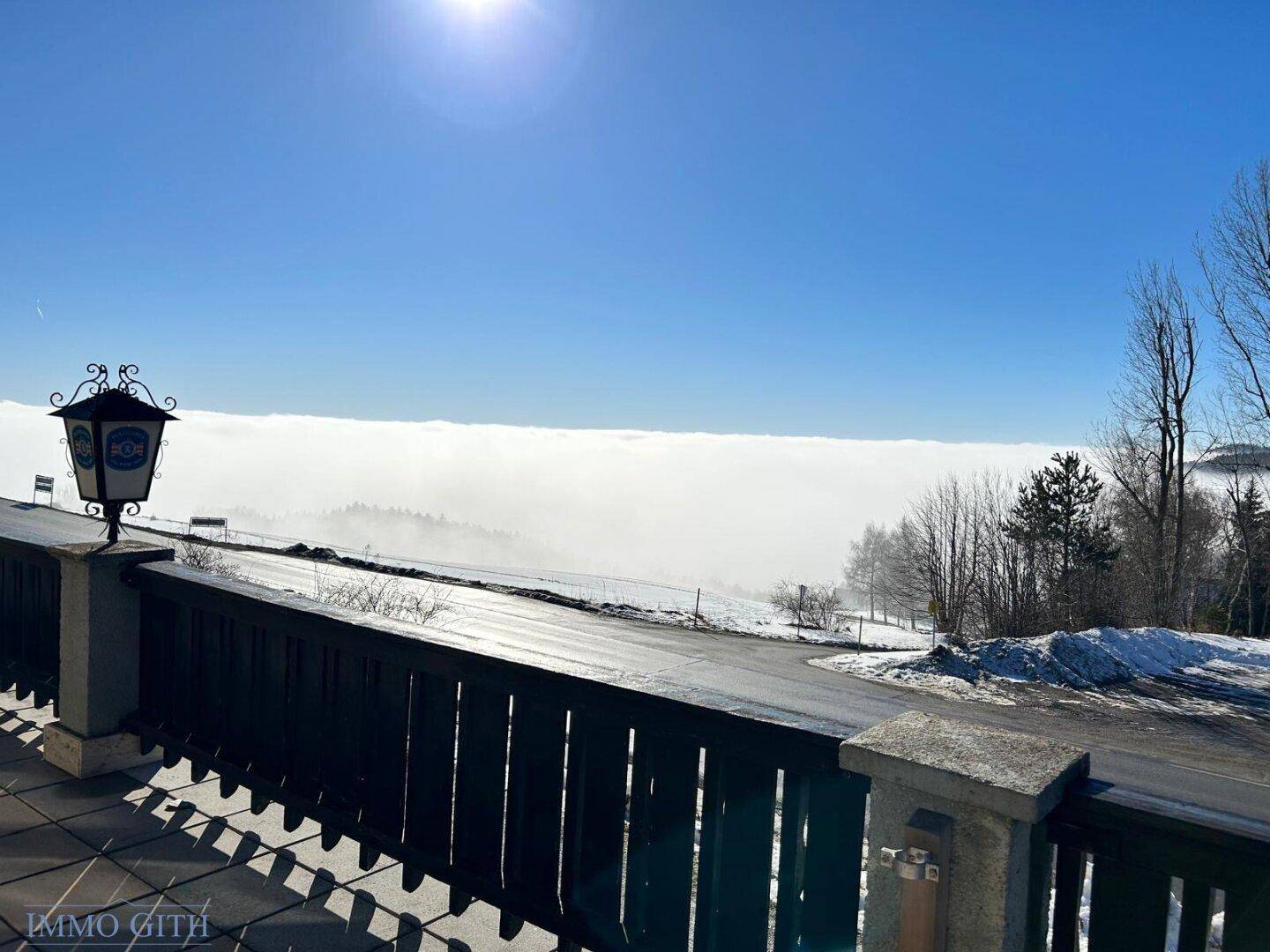 Atemberaubender Ausblick von einer Terrasse über ein Wolkenmeer und verschneite Landschaft an einem sonnigen Tag.