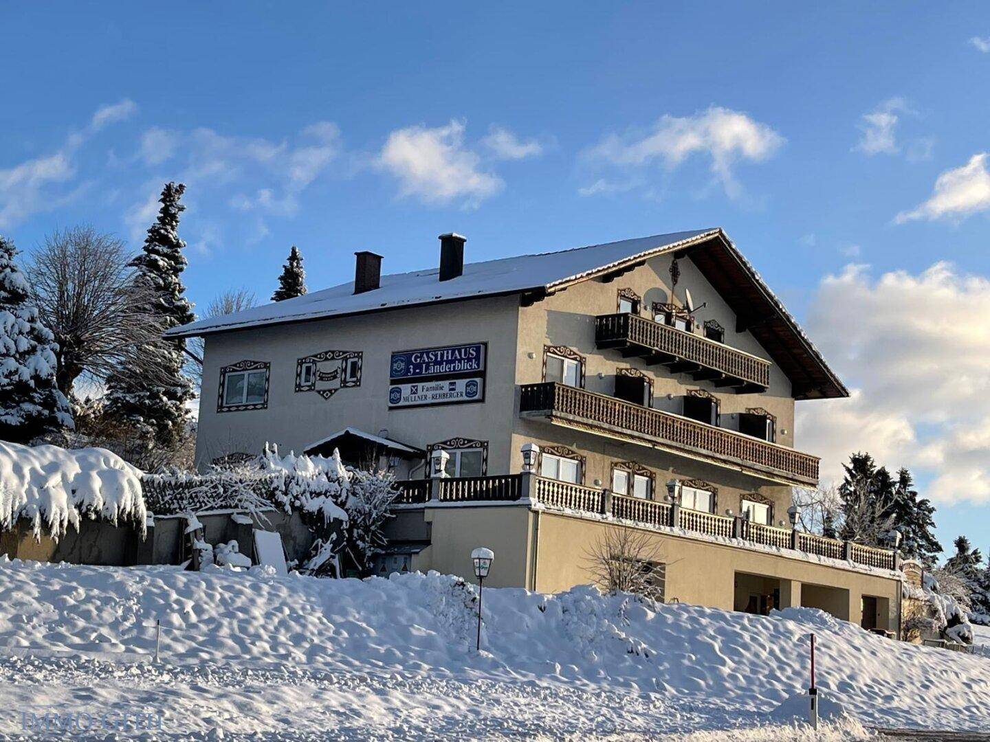Traditionelles Gasthaus in verschneiter Winterlandschaft mit Balkonen und einem Giebeldach unter blauem Himmel.