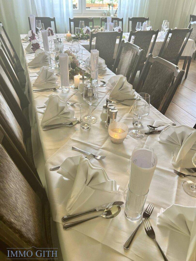 Langer, elegant gedeckter Esstisch mit weißen Tischdecken, Kerzen und Besteck für eine Feierlichkeit.