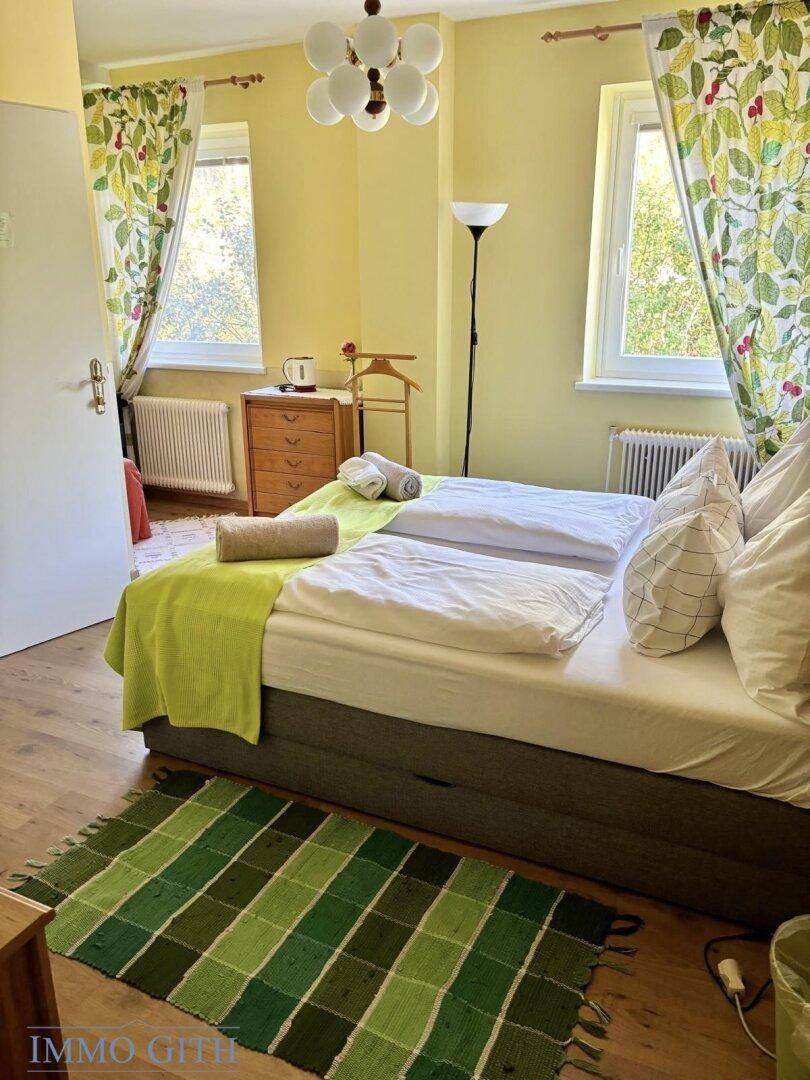 Helles Schlafzimmer mit Doppelbett, zwei Fenstern und einem grünen Teppich, freundlich gestaltet.