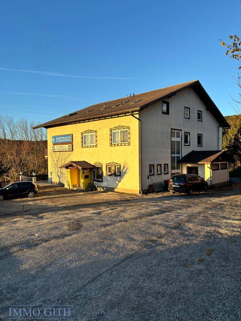 Gasthaus mit großem Parkplatz vor dem Gebäude, umgeben von Bäumen unter strahlend blauem Himmel.