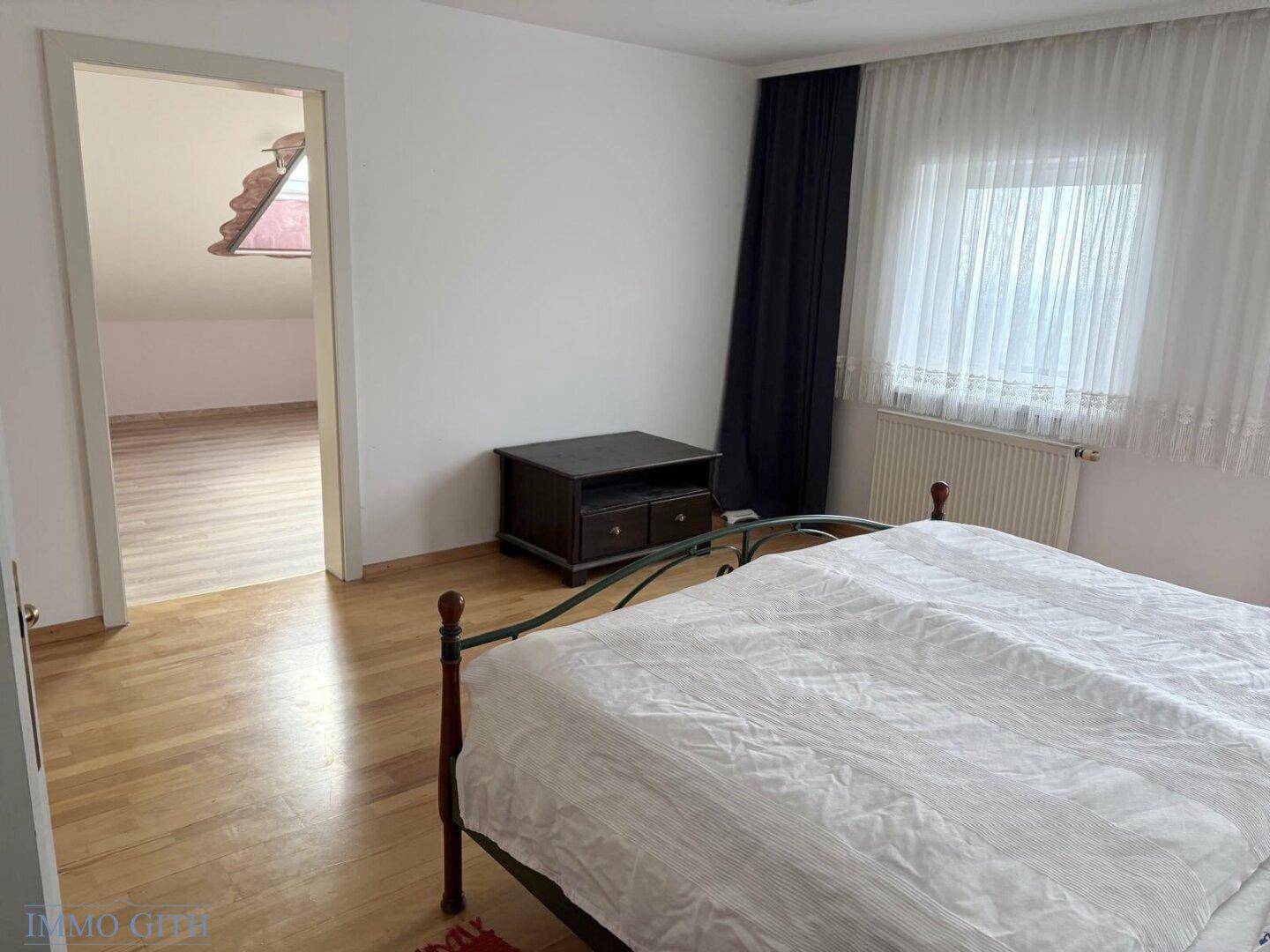 Helles Schlafzimmer mit Doppelbett, Fenster und Zugang zu einem weiteren Raum mit Holzboden.