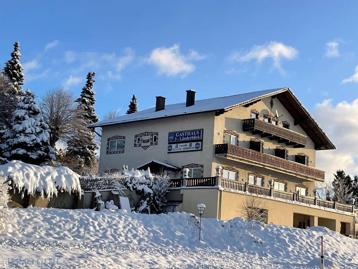 Traditionelles Gasthaus in verschneiter Winterlandschaft mit Balkonen und einem Giebeldach unter blauem Himmel.