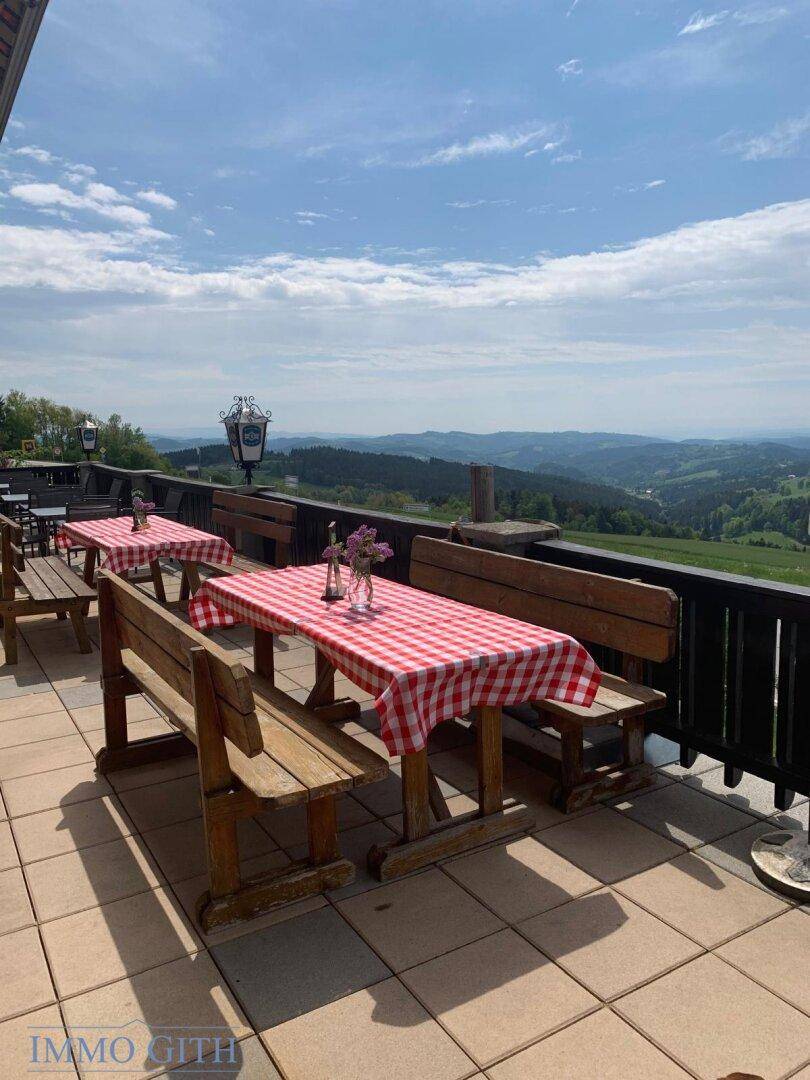 Gemütliche Außenterrasse mit Holztischen und Bänken, rot-weiß karierten Tischdecken und weitem Talblick.