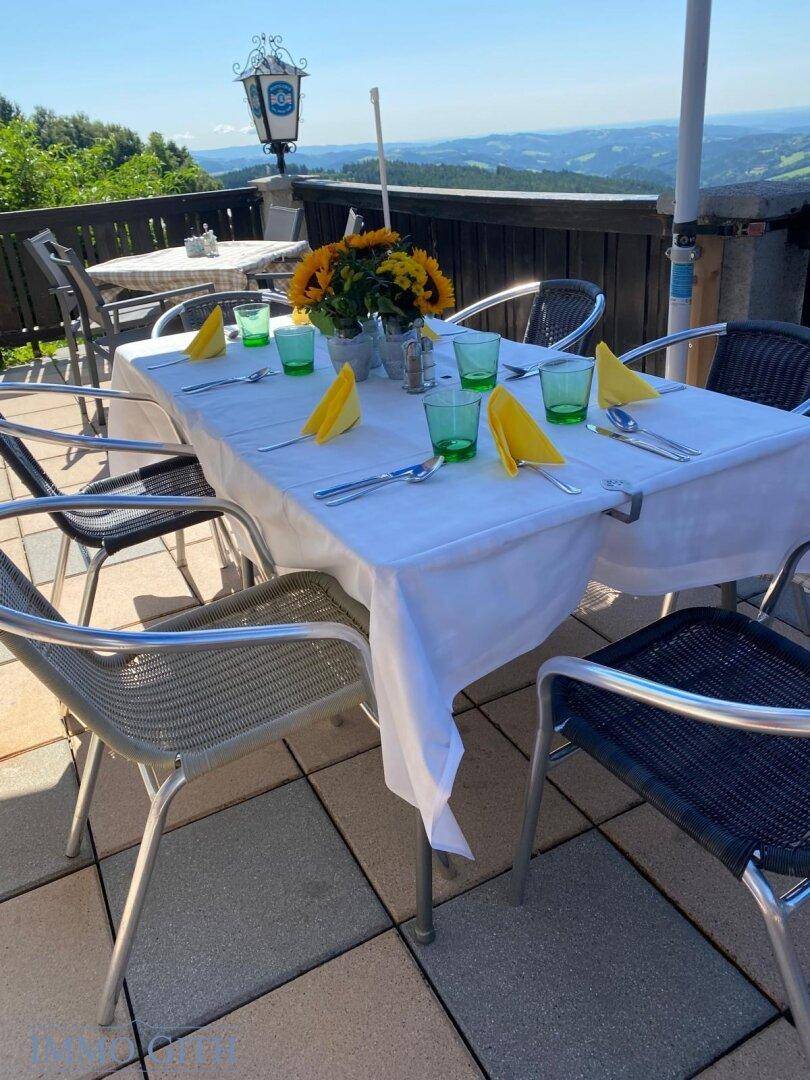 Gedeckter Terrassentisch mit weißen Tischdecken, gelben Servietten und grünen Gläsern, mit Panoramablick.