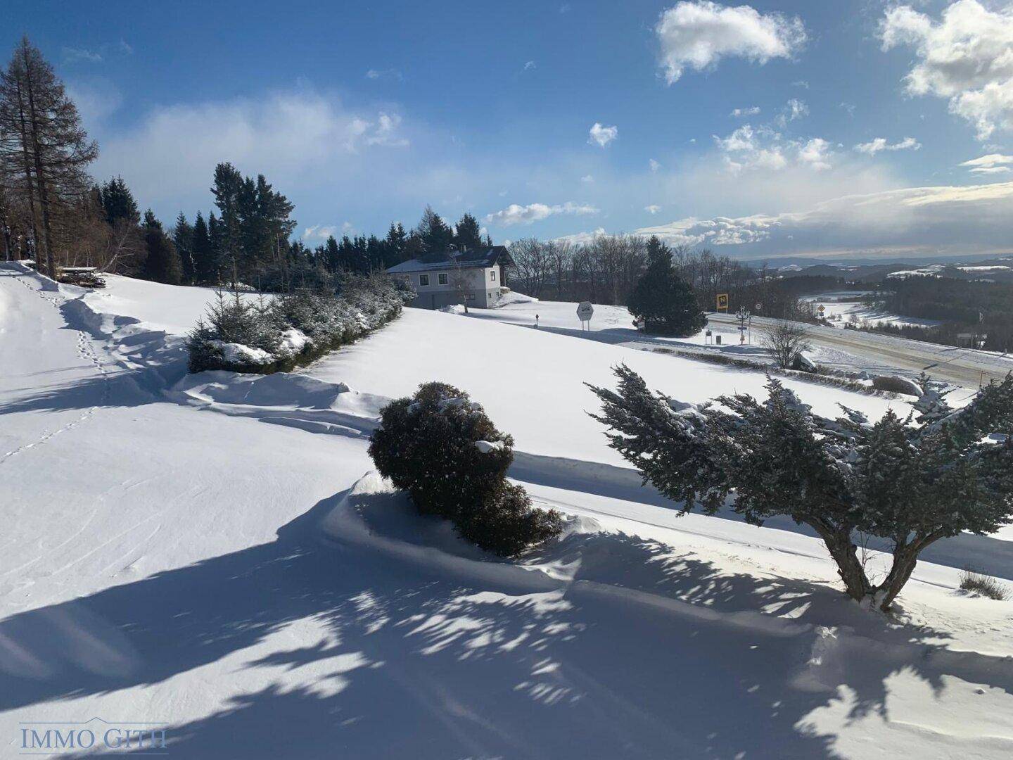 Weite, verschneite Landschaft mit einem Gebäude in der Ferne und Bäumen unter blauem Himmel.