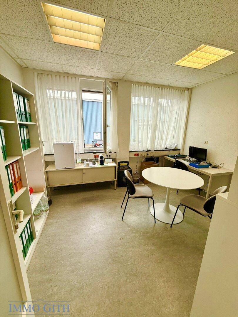 Helles Büro mit einem kleinen Besprechungstisch, Regalen voller Aktenordner und großen Fenstern.