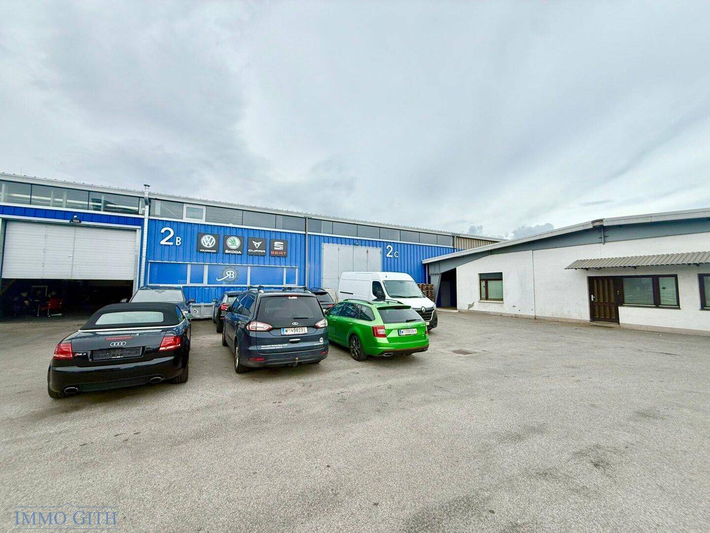 Außenansicht eines Gewerbekomplexes mit großem Asphaltparkplatz und mehreren geparkten Fahrzeugen vor blauer Fassade.