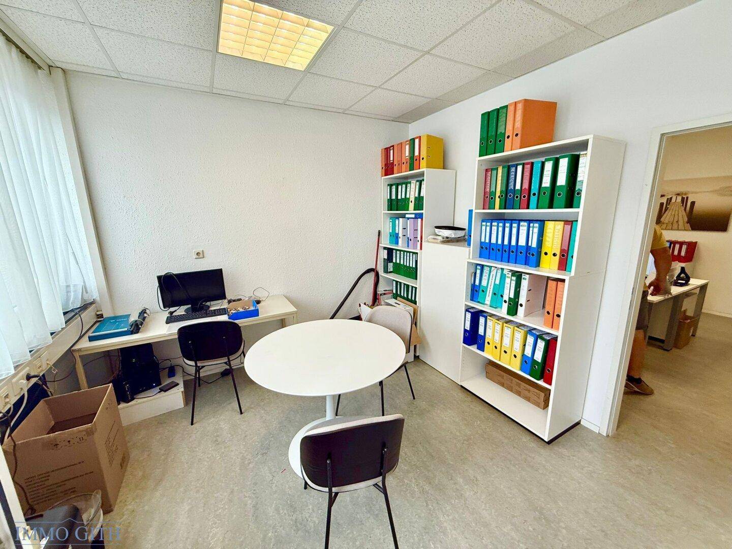 Büro mit mehreren Regalen voller bunter Aktenordner, einem kleinen Tisch und Stühlen.