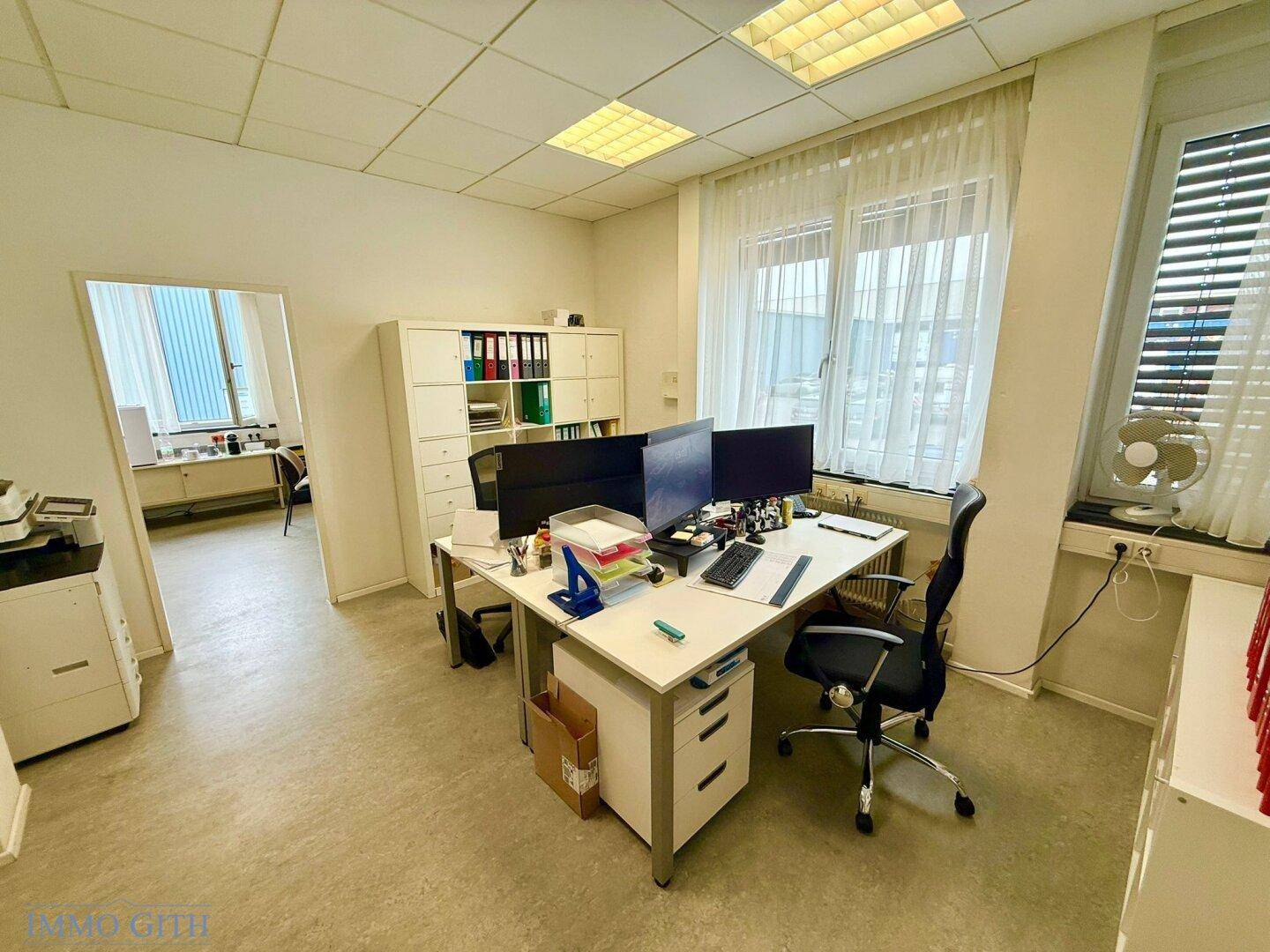 Helles Büro mit einem Arbeitsplatz, Computern und einem offenen Durchgang zu einem weiteren Raum.