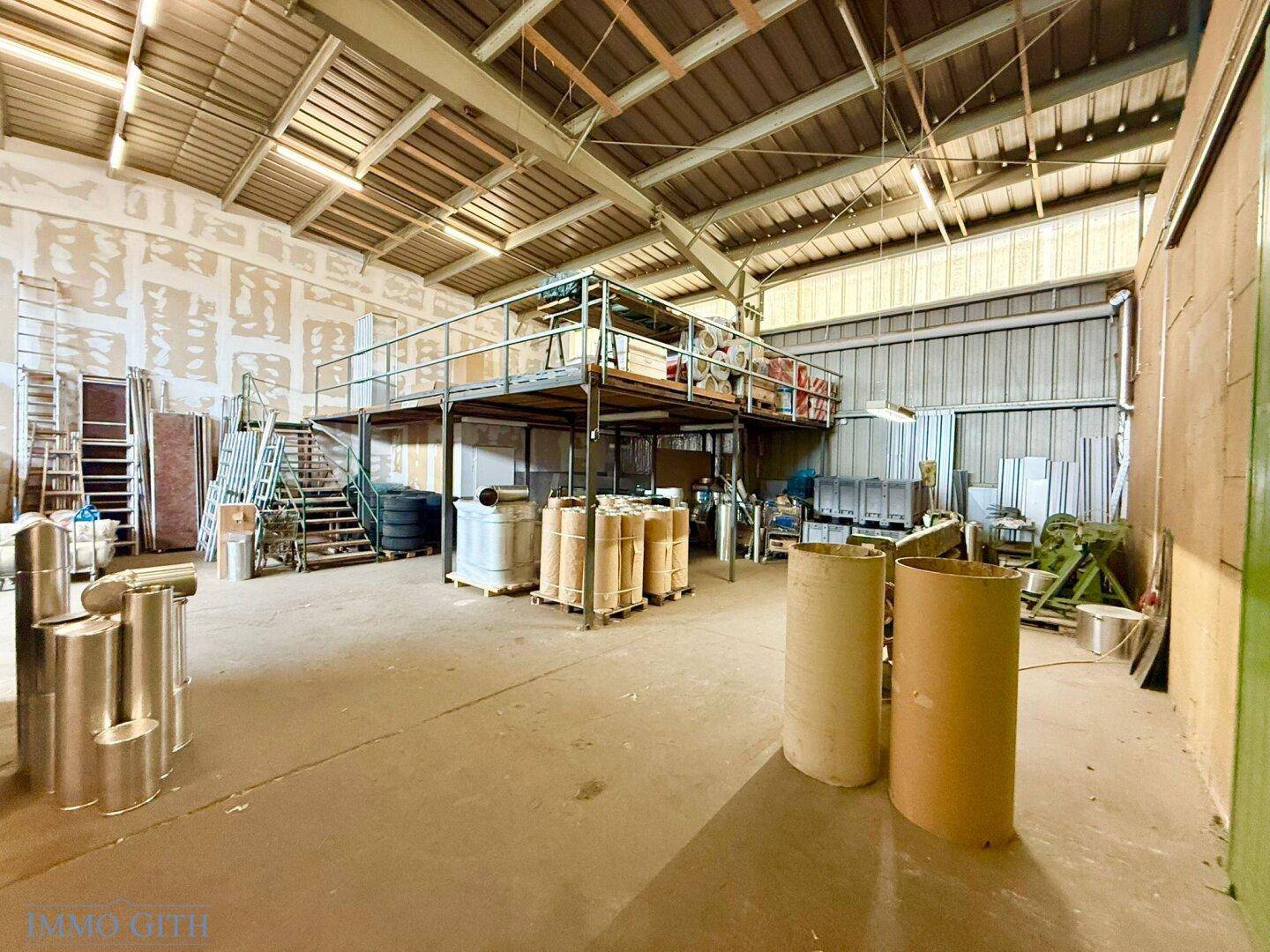 Geräumige Lagerhalle mit hoher Decke, Zwischengeschoss und gestapelten Materialien für industrielle Nutzung.