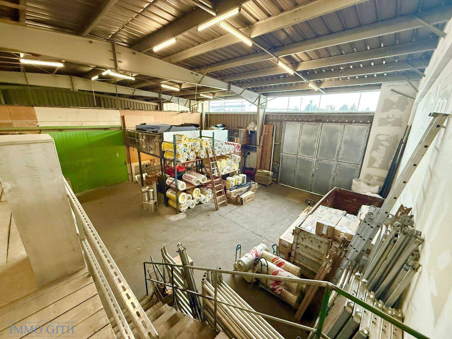 Blick von oben in eine Lagerhalle mit gestapelten Materialien, einer großen Rolltür und einer Leiter.