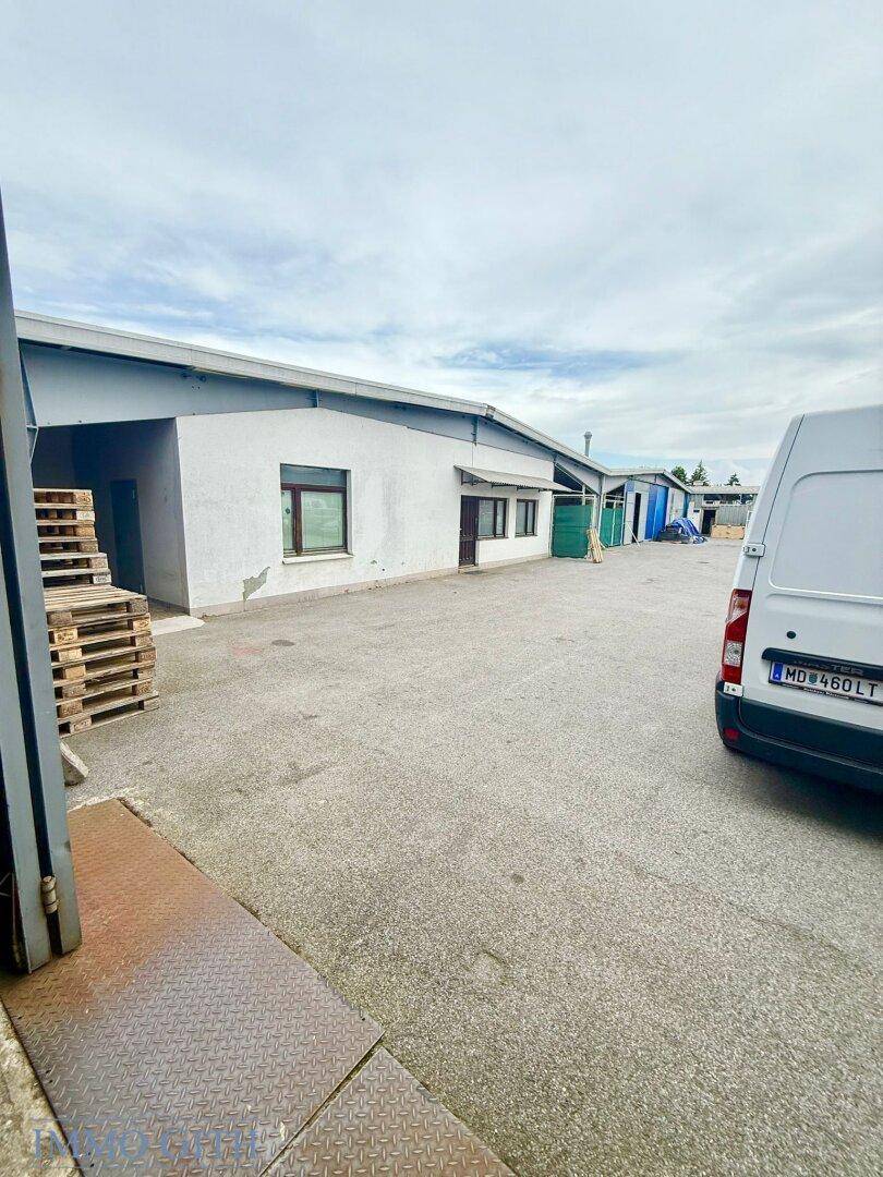 Außenansicht eines Gewerbegebäudes mit weißer Fassade und großem asphaltierten Vorplatz, ein Lieferwagen parkt.