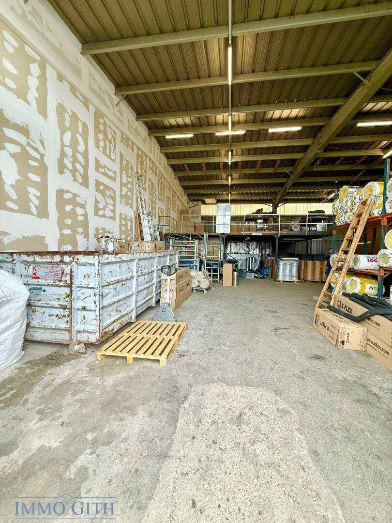 Großer Lagerraum mit Betonboden, hohen Decken und einer Zwischenebene, gefüllt mit Materialien und einem Container.