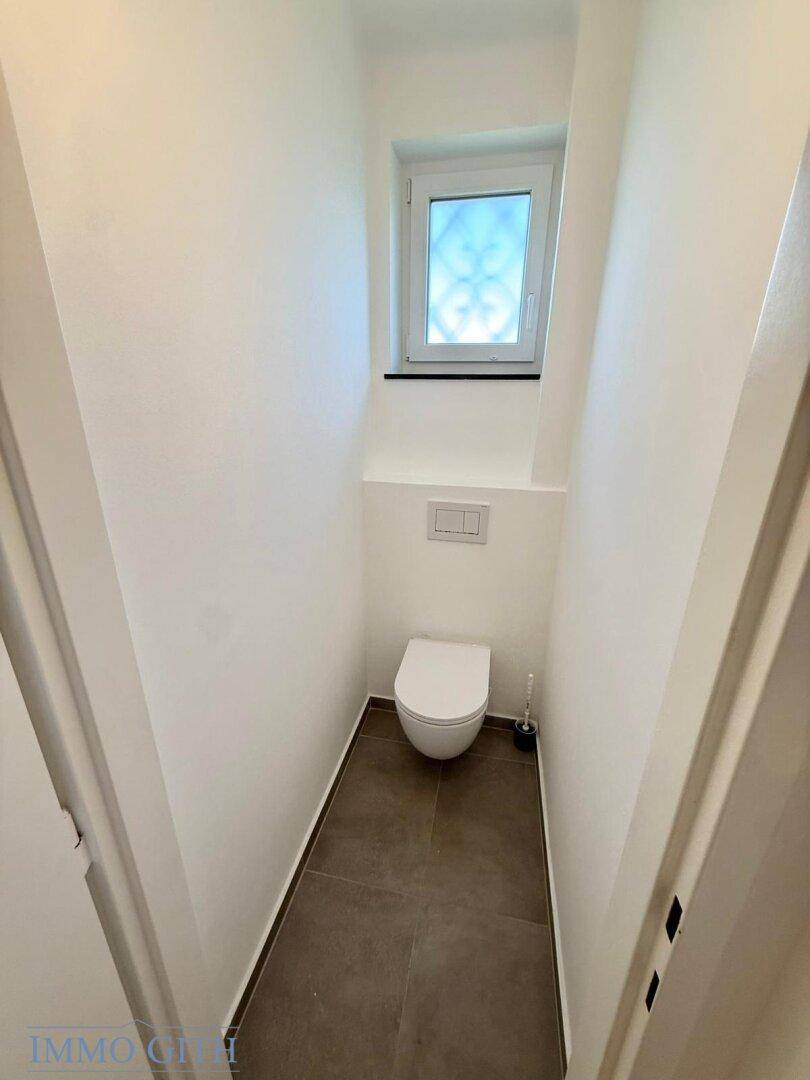 Separates WC mit wandhängender Toilette und kleinem Fenster, modern und funktional gestaltet.