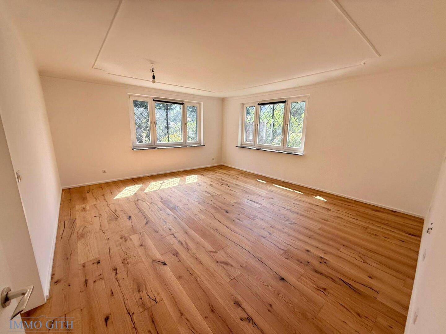 Helles, leeres Wohnzimmer mit neuem Holzboden und zwei Fenstern, die viel Tageslicht hereinlassen.