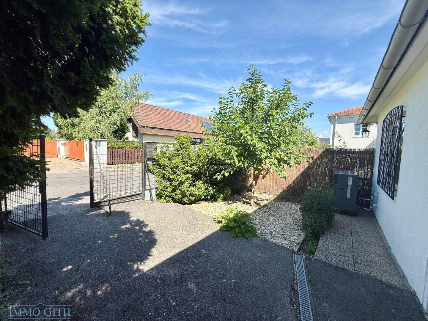 Sonniger Gartenbereich mit Bäumen, Kies und Zaun, bietet Privatsphäre und Erholung.