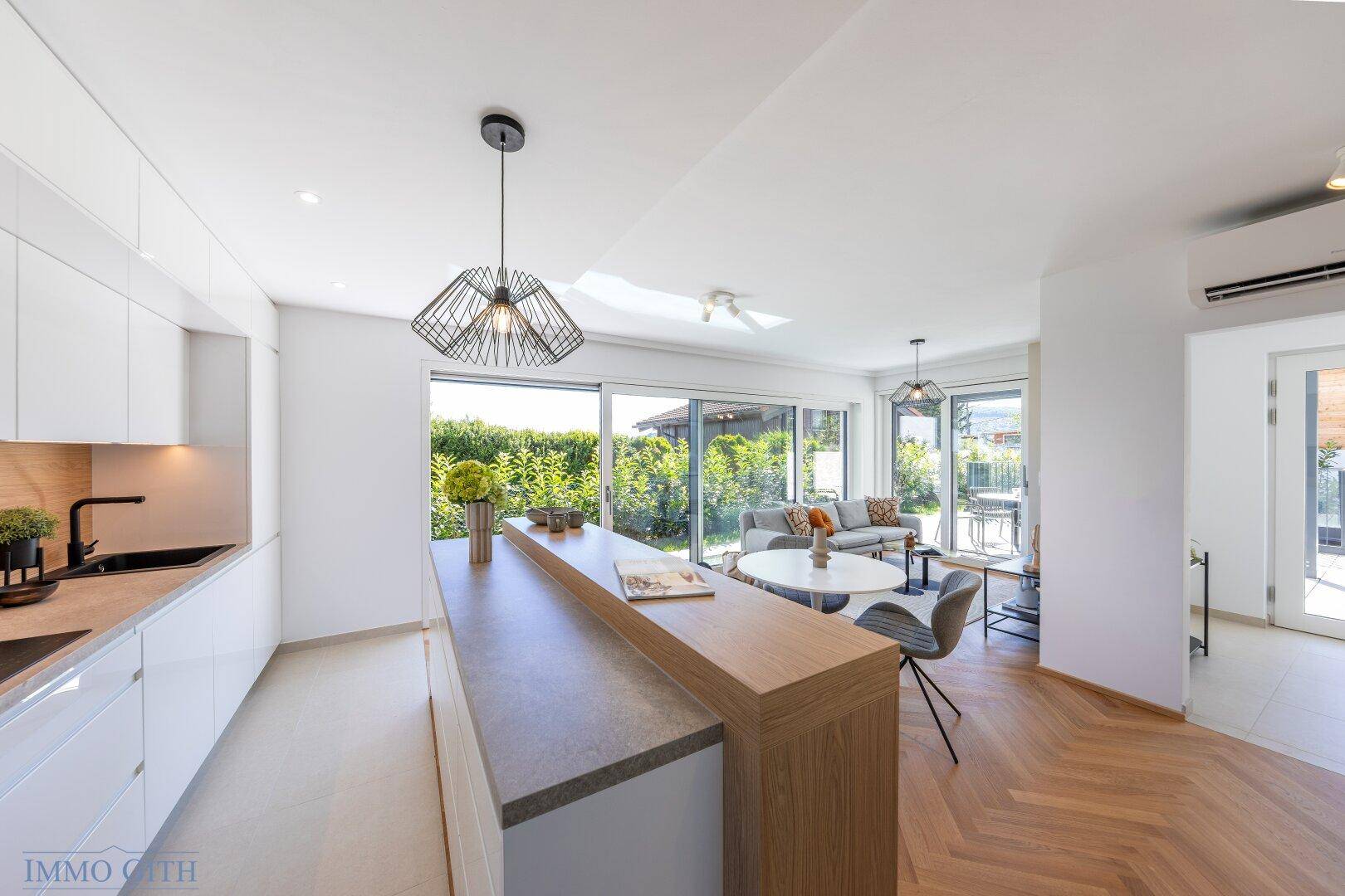 Moderne weiße Küche mit Kochinsel und Frühstücksbar, offen zum Wohnbereich mit Gartenblick.