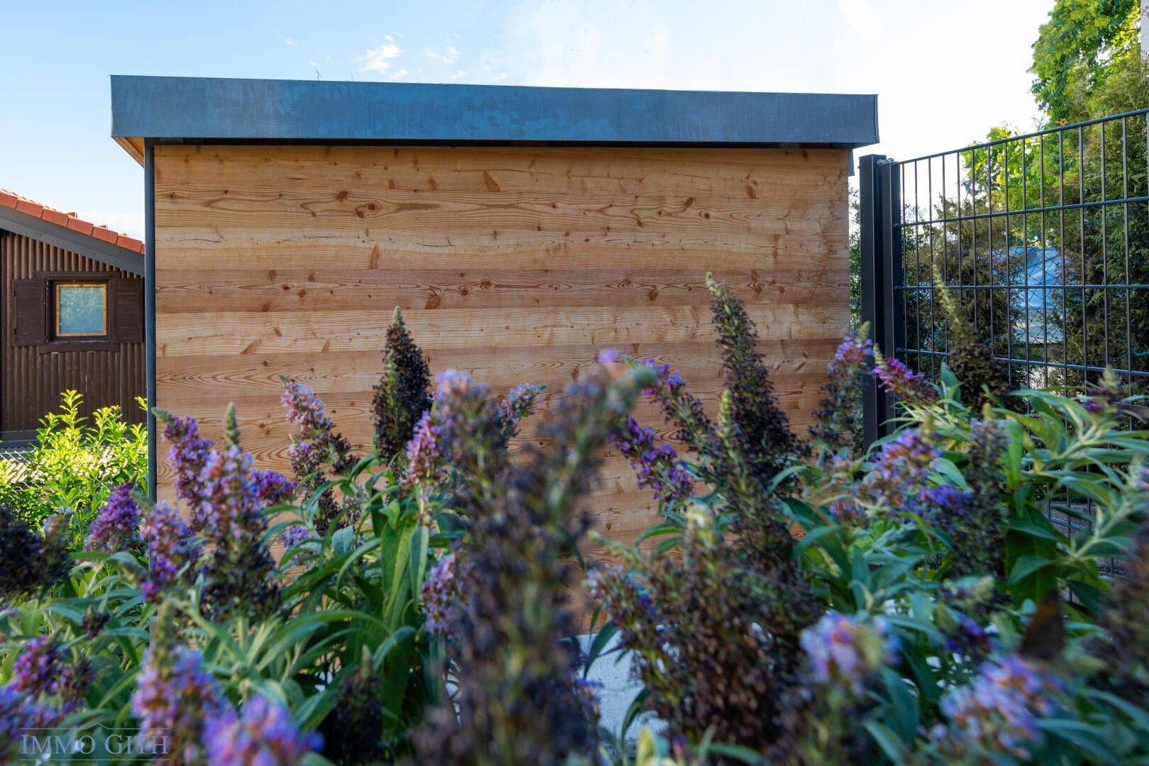 Holzverkleidete Außenwand umgeben von blühenden Pflanzen in einem gepflegten Gartenbereich.