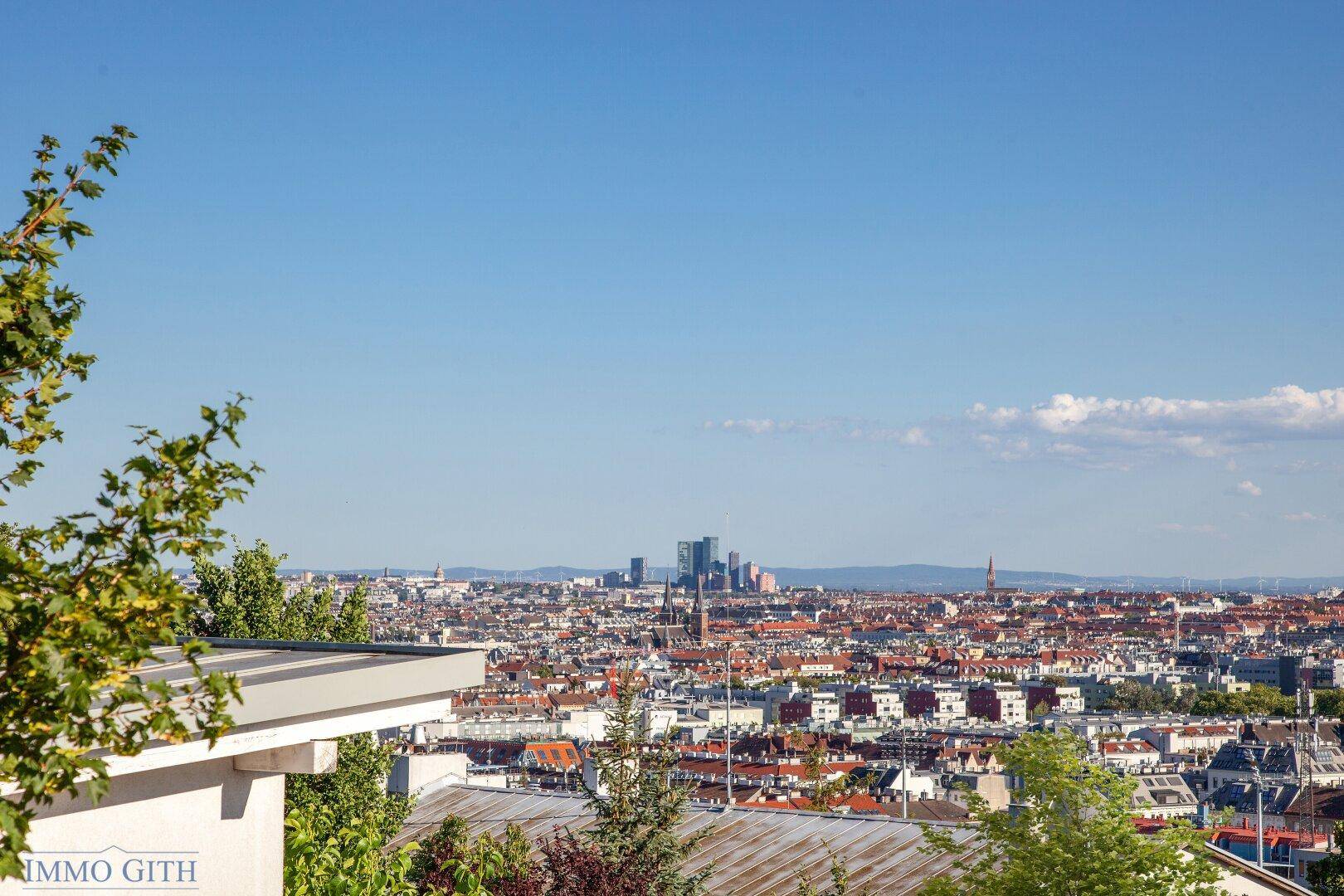 Panoramablick auf die Stadtlandschaft unter blauem Himmel, umgeben von grünen Bäumen.