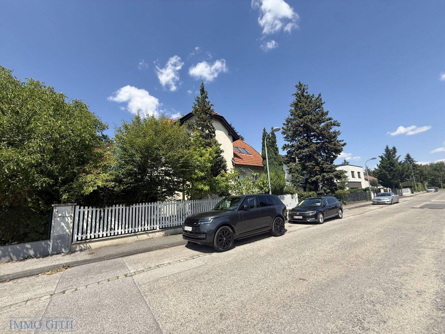 Weitläufige Ansicht des Hauses von der Straße mit Bäumen und geparkten Fahrzeugen.