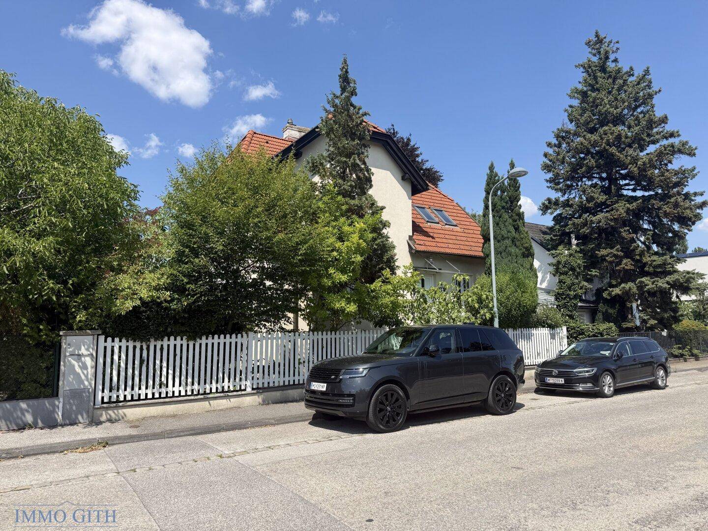 Das Haus von der Straße aus gesehen, umgeben von viel Grün und mit einem weißen Zaun.