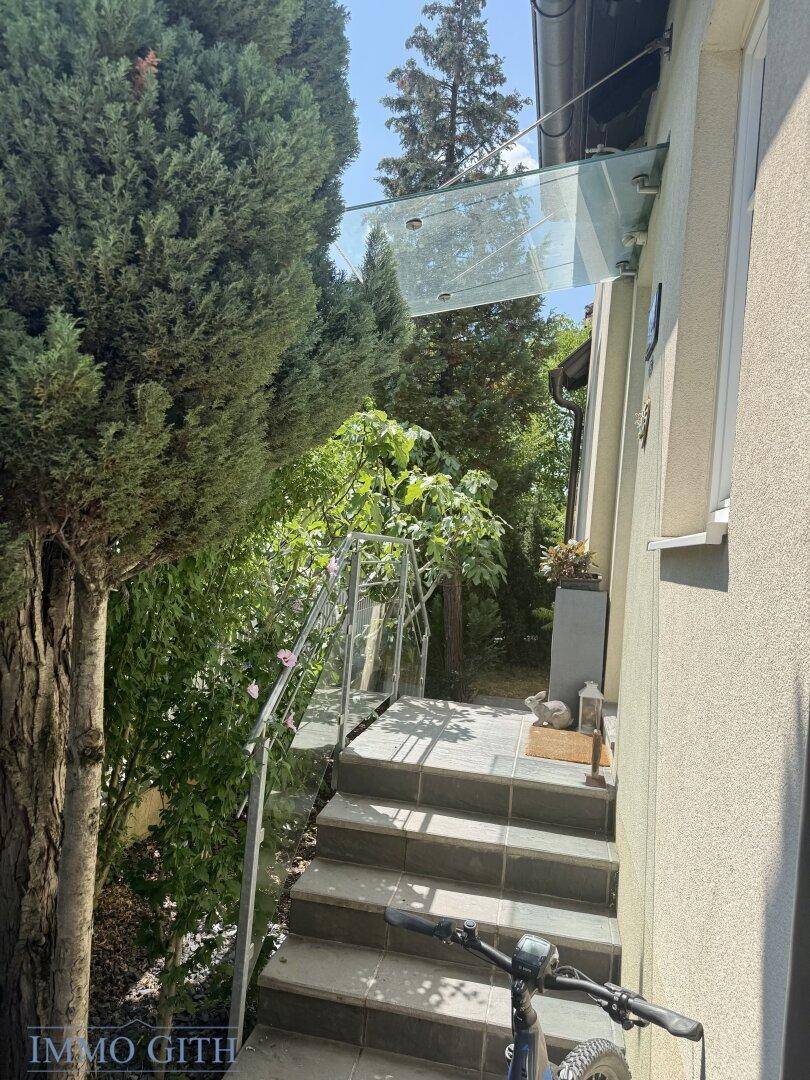 Eingangstreppe mit moderner Glasüberdachung, umgeben von üppigem Grün.
