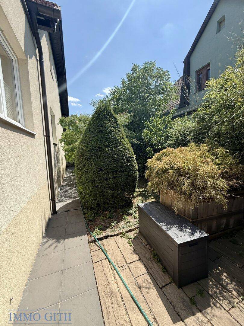 Schmaler Weg zwischen Gebäuden, umgeben von dichtem Grün und hohen Hecken.