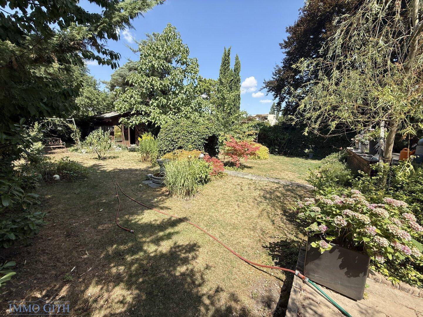 Weitläufiger Garten mit Rasenfläche, vielfältiger Bepflanzung und einem kleinen Gartenhaus im Hintergrund.