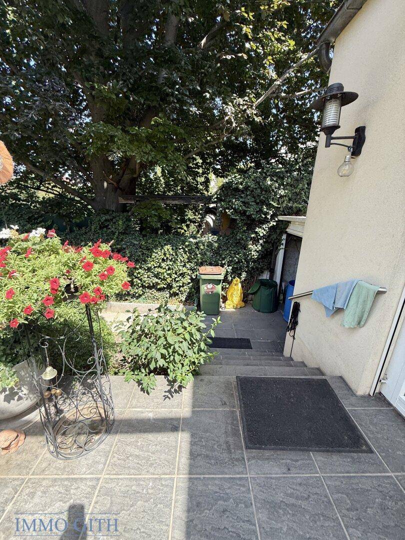Gefliester Außenbereich mit Treppe, Wandleuchte und blühenden Topfpflanzen.